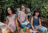 Three girls sit on a wooden table in the sun—one in a pink butterfly reversible dress, one in a pink butterfly tank top, and one in a blue leopard tank top