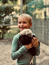A blonde boy smiles wearing the sage green long pyjamas, whilst holding a chicken happily.