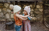 Two young girls in cosy knit hats smile as they hold each other’s arms. One wears a super soft pink butterfly dress, the other an orange plush dinosaur vest.