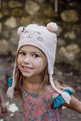 A young girl wearing a pink butterfly-print dress with ruffled sleeves and a knitted cat-ear hat smiles against a rustic stone wall. Her soft, sensory-friendly clothing is designed for comfort and ease.