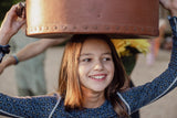 A smiling girl with long black hair in a soft leopard pyjamas holding an old leather hat box on her head.