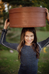 A smiling girl with long black hair in a soft leopard pyjamas holding an old leather hat box on her head.