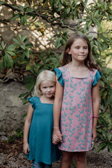 Two girls in reversible butterfly dresses face the camera, now wearing the opposite sides—one in teal blue, the other in pink with butterflies.