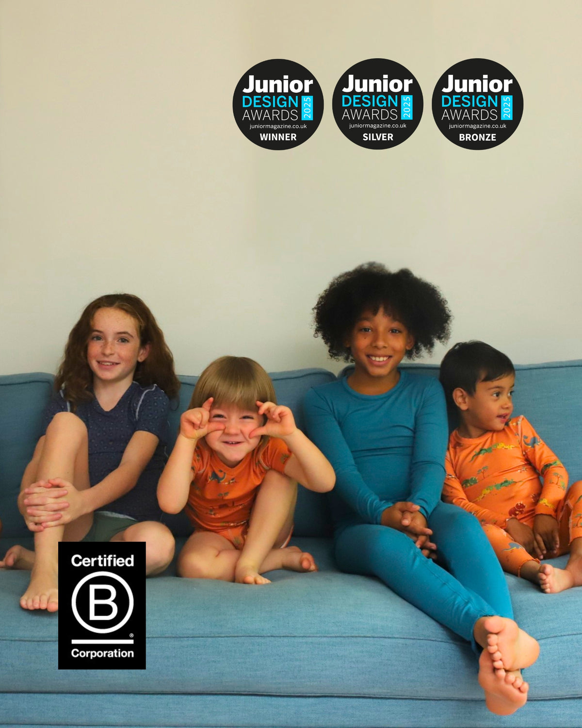 Four children sitting on a blue couch in matching orange and blue pajamas, with three circular 'Junior Design Awards' badges at the top and a 'Certified B Corporation' logo at the bottom left.