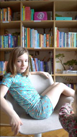 Girl spins on chair wearing the sensory friendly aqua blue fruit splash short pyjamas, with a bookcase of the bedroom in the background