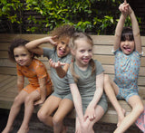 Four girls are sat on a garden bench, happily smiling wearing soft sensory friendly short pyjamas in sage green, aqua blue and orange dinosaur patterns.