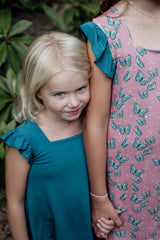 A young blonde-haired girl in a teal ruffled-sleeve dress leans against an older child wearing a pink dress adorned with teal butterflies. The two hold hands, showcasing the softness and sensory-friendly design of the fabric. The natural outdoor background adds to the warmth and comfort of the moment.